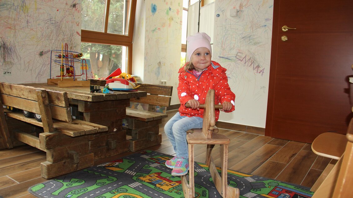 Ein Mädchen sitzt auf einem Holzspielzeug in einem bunten Spielzimmer. Die Wände sind mit Kritzeleien dekoriert, und es gibt Spielzeug und Möbel aus Holz. | © Riedenbauer