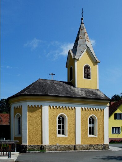 A small yellow chapel with a pointed roof and windows in the Gothic style. It stands in a tranquil setting with a blue sky in the background. | © MG Eibiswald