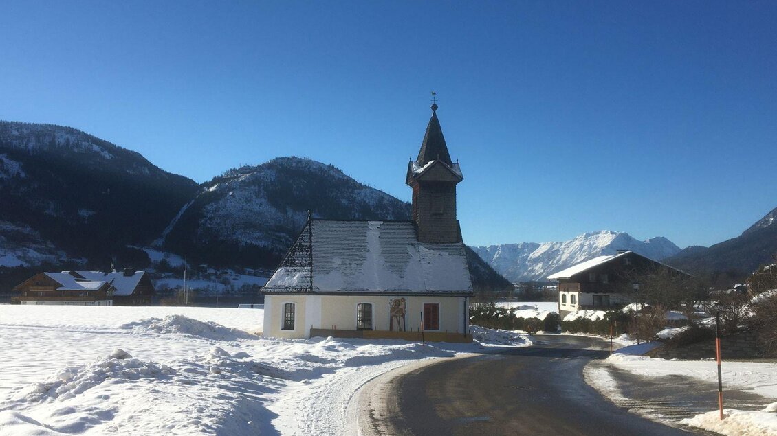 Eine malerische Kirche in einer verschneiten Landschaft. Im Hintergrund sind Berge und ein blauer Himmel sichtbar.