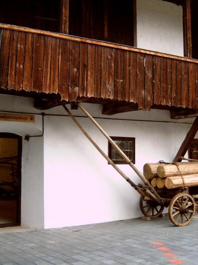 Ein traditionelles Gebäude mit einer Holzfassade und einem großen Fenster. Vor dem Gebäude steht ein Wagen mit gestapelten Holzstämmen. | © Gemeinde Hirschegg-Pack