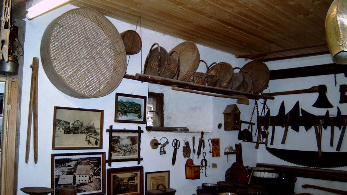 Ein rustikales Zimmer mit alten Werkzeugen und Holzgegenständen. An den Wänden hängen Bilder und Körbe, die die traditionelle Handwerkskunst zeigen. | © Gemeinde Hirschegg-Pack