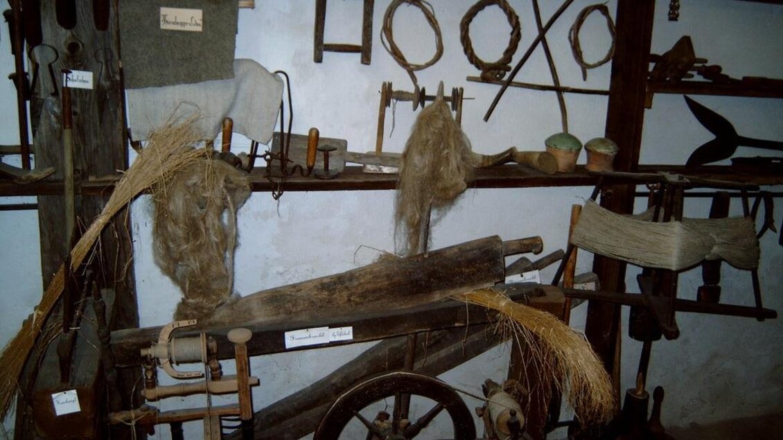 Eine Sammlung von historischen Handwerkszeugen und Maschinen in einem Museum. Auf Regalen sind verschiedene Materialien und Geräte für die Textilproduktion zu sehen. | © Gemeinde Hirschegg-Pack