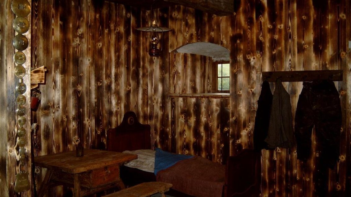 Ein rustikales Zimmer mit Holzvertäfelung und einer einfachen Einrichtung. Es gibt ein Bett, einen Tisch und einige Kleidungsstücke an der Wand. | © Gemeinde Hirschegg-Pack