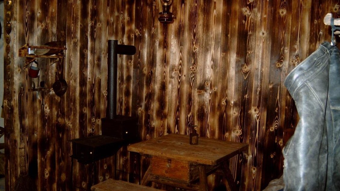 Ein rustikales Zimmer mit einer Holzwand und einem alten Tisch. Auf der linken Seite hängen ein Stuhl und eine Lampe. | © Gemeinde Hirschegg-Pack