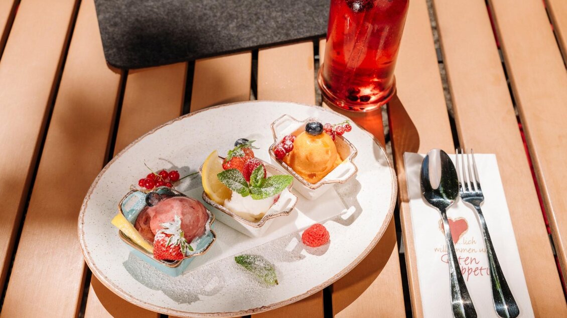 Ein Teller mit drei verschiedenen Eissorten, garniert mit Beeren und frischen Minzblättern. Daneben steht ein Getränk in einer Flasche und Besteck auf einem Tisch. | © Gasthaus Stöckl