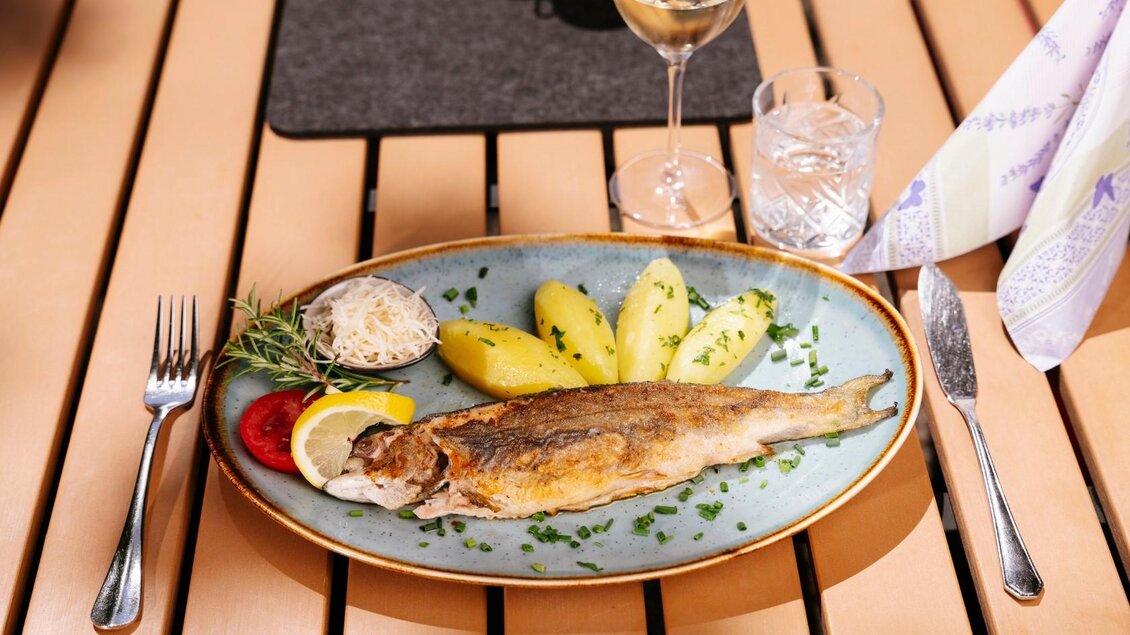 Ein schöner Teller mit gebratenem Fisch, Beilagen von Kartoffeln und Salat. Dazu ein Glas Weißwein auf einem Holz Tisch. | © Gasthaus Stöckl
