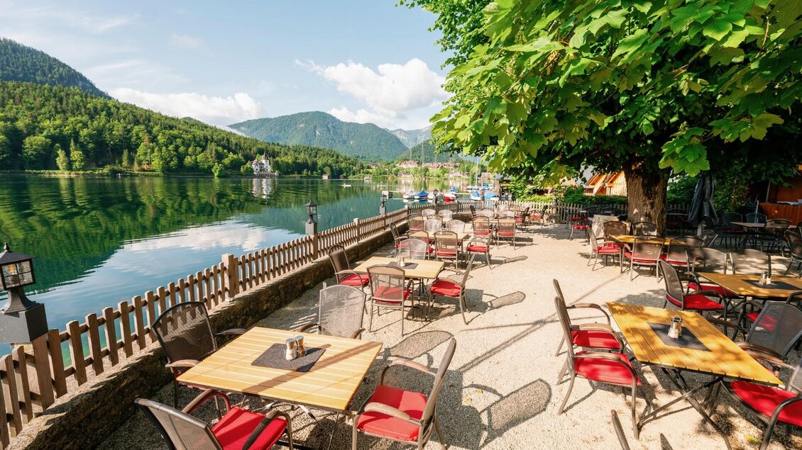 Ein einladendes Café am Ufer eines ruhigen Sees mit Tischen und Stühlen in der Sonne. Im Hintergrund sind grüne Berge und ein klarer Himmel zu sehen. | © Gasthaus Stöckl