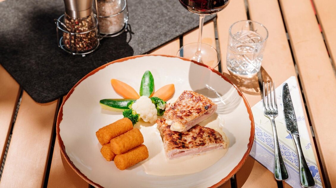 Ein Teller mit einem Hauptgericht aus Fleisch, serviert mit Gemüse und Kartoffelgraten. Dazu ein Glas Wasser und ein Glas Rotwein. | © Gasthaus Stöckl