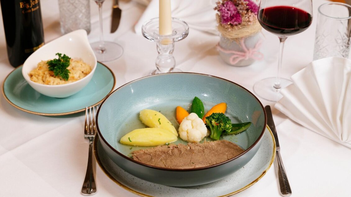 Ein elegantes Gericht mit zartem Fleisch, Gemüse und Kartoffeln auf einem Tisch gedeckt mit Rotwein und einer Kerze. Im Hintergrund ist eine Schüssel mit Beilagen zu sehen. | © Gasthaus Stöckl