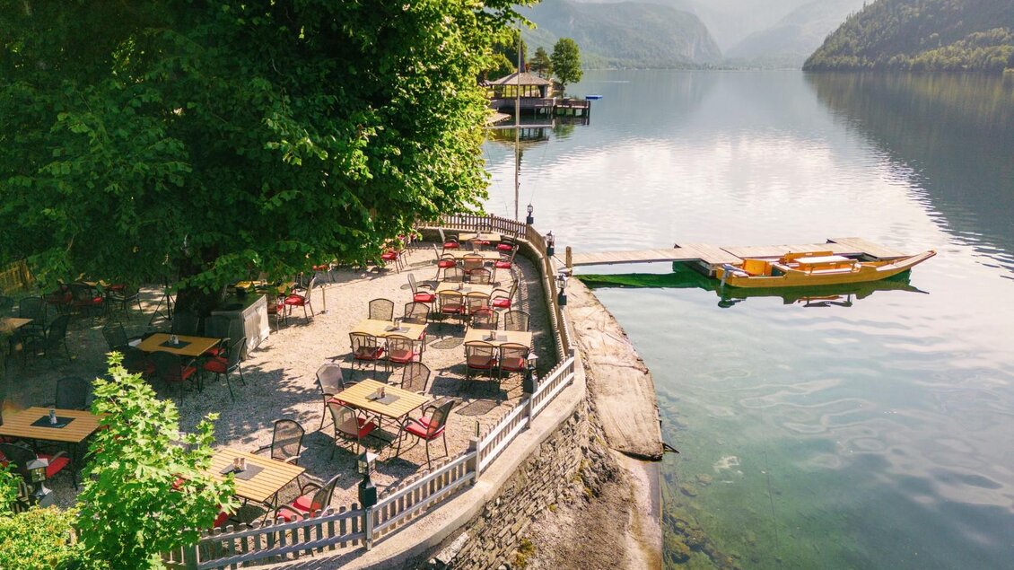Ein malerisches Restaurant am Wasser mit Tischen und Stühlen im Freien. Im Hintergrund sind Berge und ein Bootssteg zu sehen. | © Gasthaus Stöckl