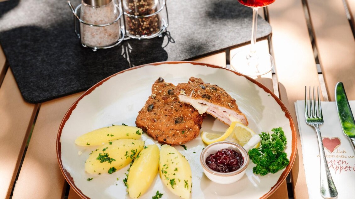 Ein Teller mit einem panierten Schnitzel, Kartoffeln und Preiselbeersoße. Dazu ein Glas Rotwein und Gewürzmühlen auf einem Tisch. | © Gasthaus Stöckl