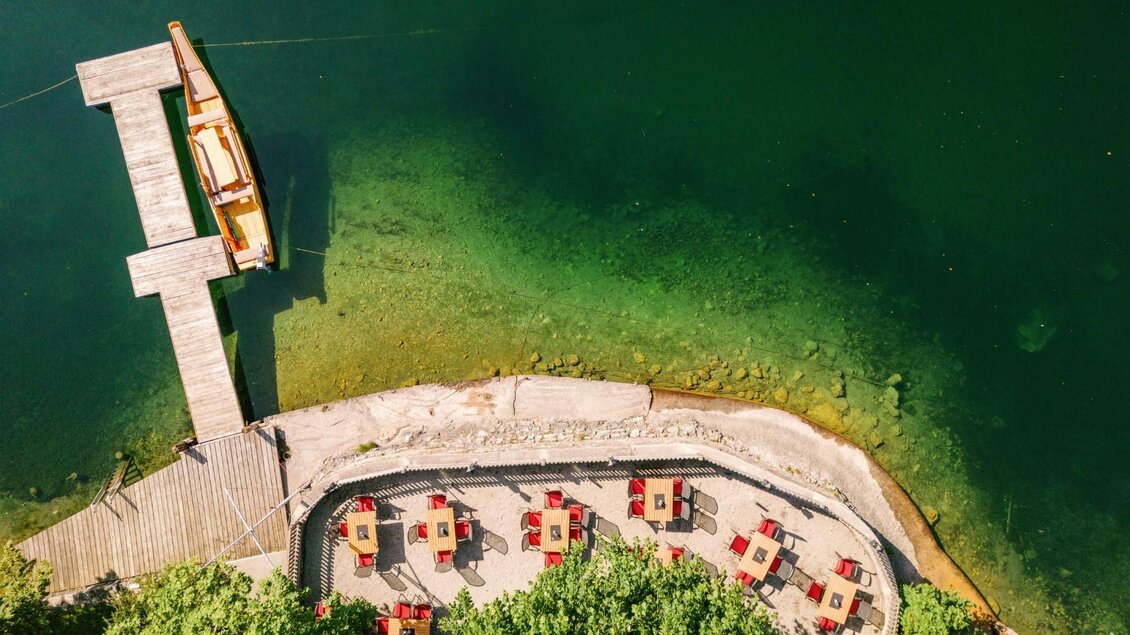 Ein ruhiger See mit einem Holzsteg und einem Boot. Auf der Uferpromenade stehen rote Tische und Stühle. | © Gasthaus Stöckl