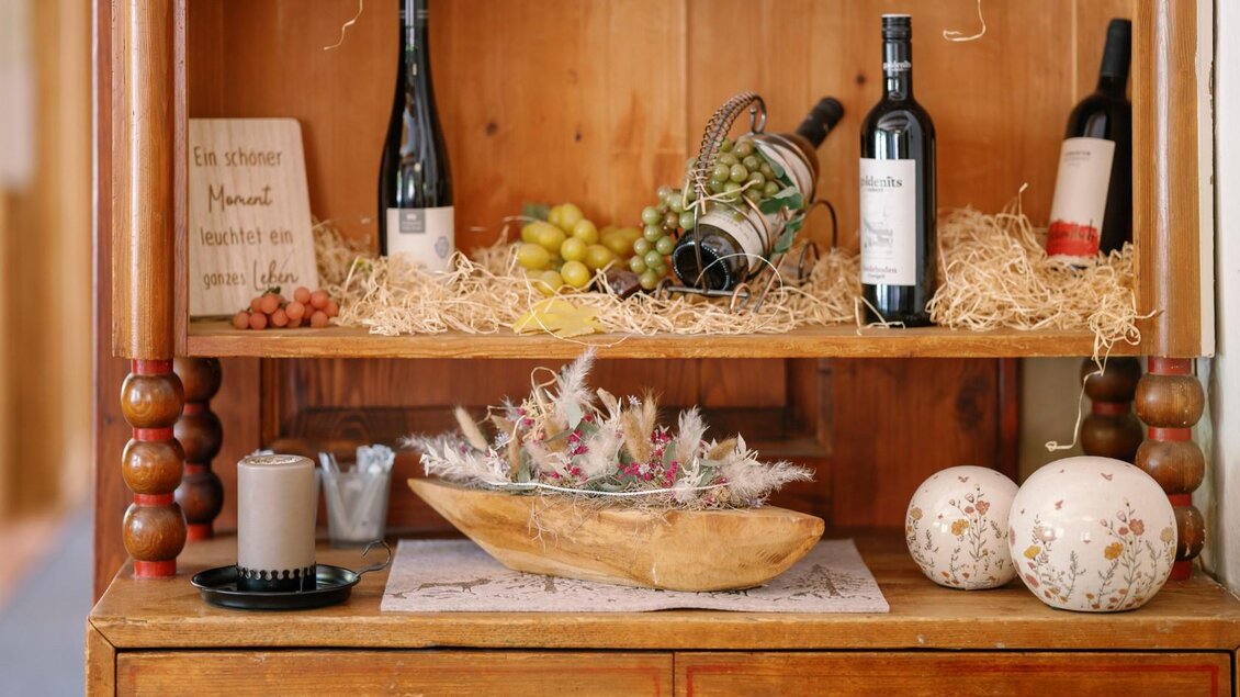 Ein rustikales Regal mit Weinflaschen, frischen Trauben und einer dekorativen Holzschale. Die warmen Farben und die natürlichen Materialien schaffen eine einladende Atmosphäre. | © Gasthaus Stöckl