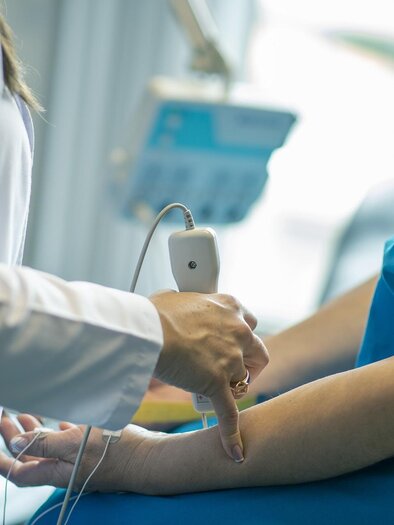 A doctor is conducting an examination with an ultrasound machine on a patient’s arm. The patient is wearing a blue hospital gown. | © Arzt