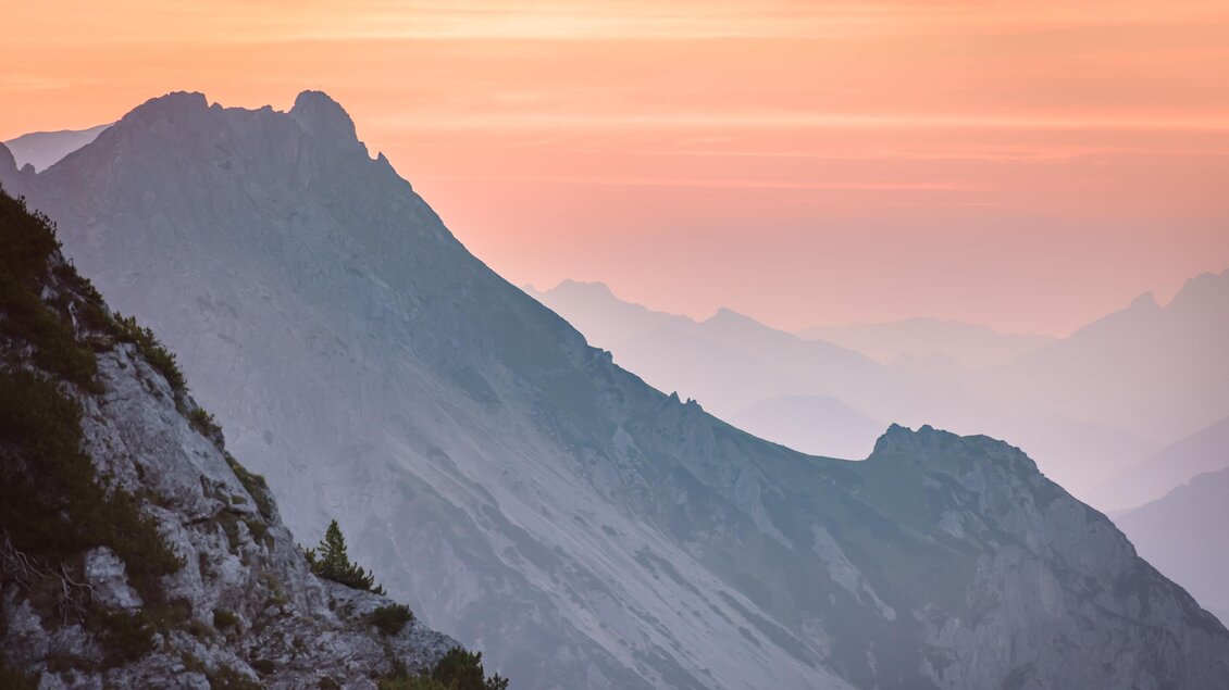 Eine malerische Berglandschaft in sanften Rosa- und Orangetönen. Die Silhouetten der Berge stechen vor dem Abendhimmel hervor. | © Christoph Huber