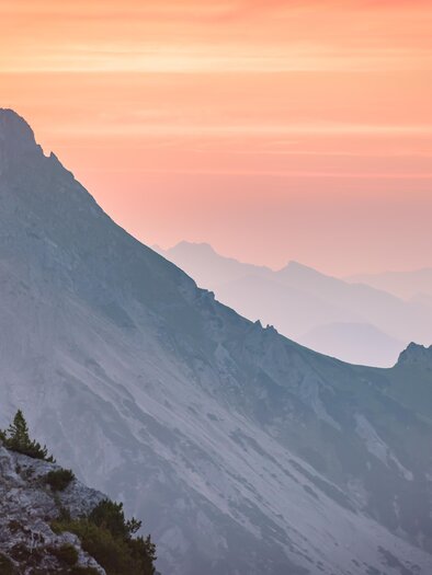 A picturesque mountain landscape in gentle shades of pink and orange. The silhouettes of the mountains stand out against the evening sky. | © Christoph Huber