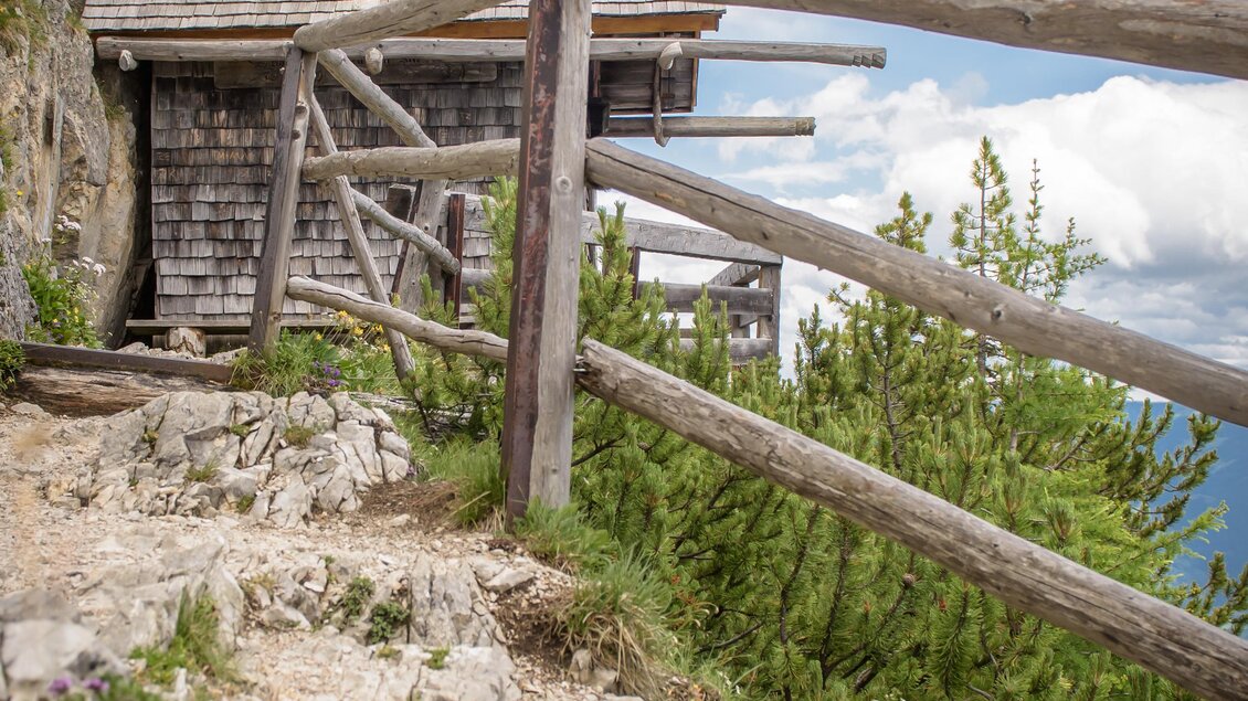 Ein malerisches Berghaus aus Holz, umgeben von Natur. Im Vordergrund blühen violette Blumen, und der Himmel ist klar und blau. | © Christoph Huber