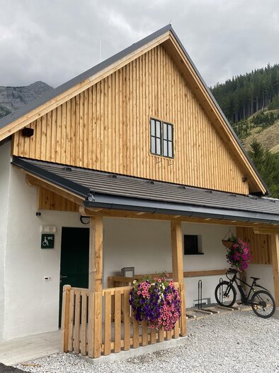 A modern wooden house with white walls and an inviting porch. In the foreground, there is a bicycle, and colorful flowers give the entrance a friendly touch. | © Zentral-Wasserversorgung Hochschwab Süd
