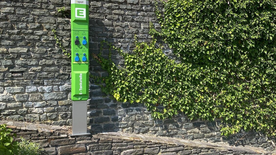 Eine Ladestation für Elektrofahrzeuge an einer Steinmauer, umgeben von Pflanzen. Im Hintergrund sind Fenster und Blumen zu sehen. | © Joachim Ruhri