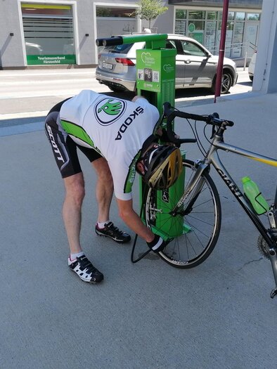 Ein Radfahrer befestigt sein Fahrrad an einer grünen Reparaturstation. Der Hintergrund zeigt eine Straße mit parkenden Autos. | © Tourismusverband Thermen-Vulkanland