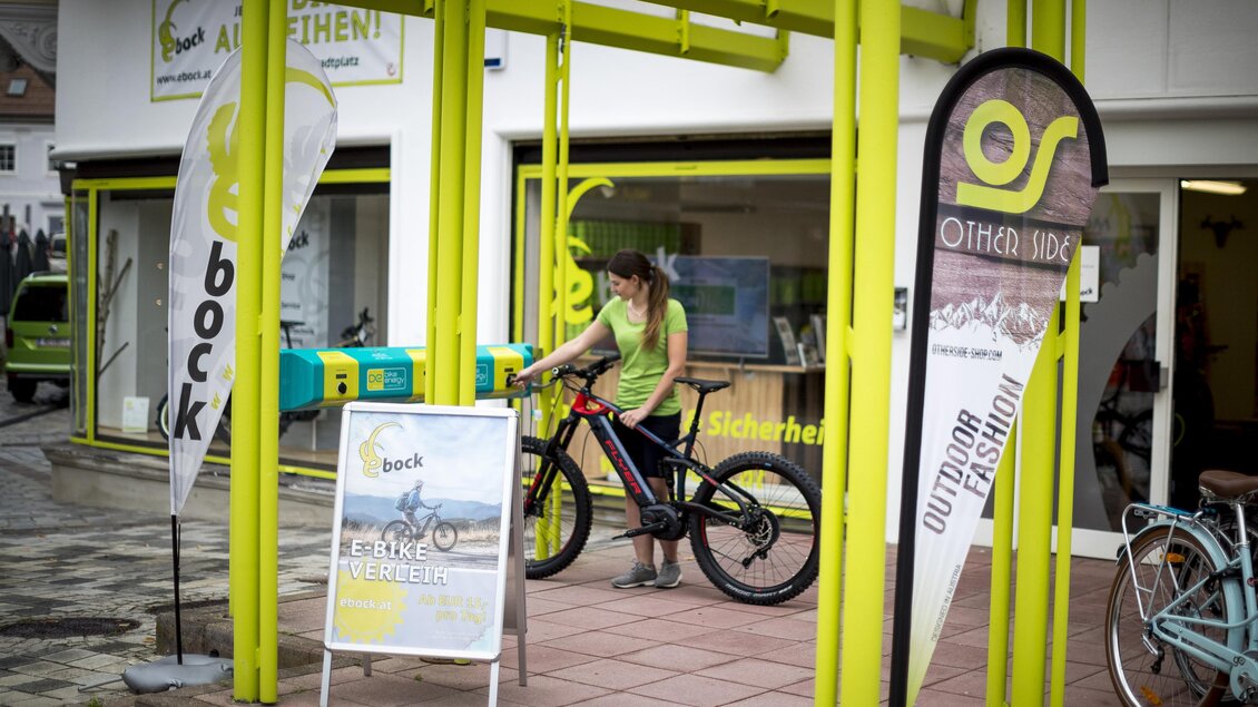 Eine Frau steht neben einem Fahrrad unter einem gelben Vordach. Im Hintergrund sind Schilder und ein Geschäft sichtbar. | © TV Hochsteiermark