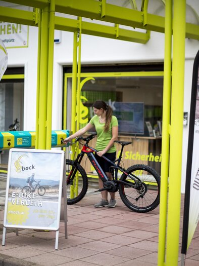 A woman stands next to a bicycle under a yellow awning. In the background, signs and a shop are visible. | © TV Hochsteiermark