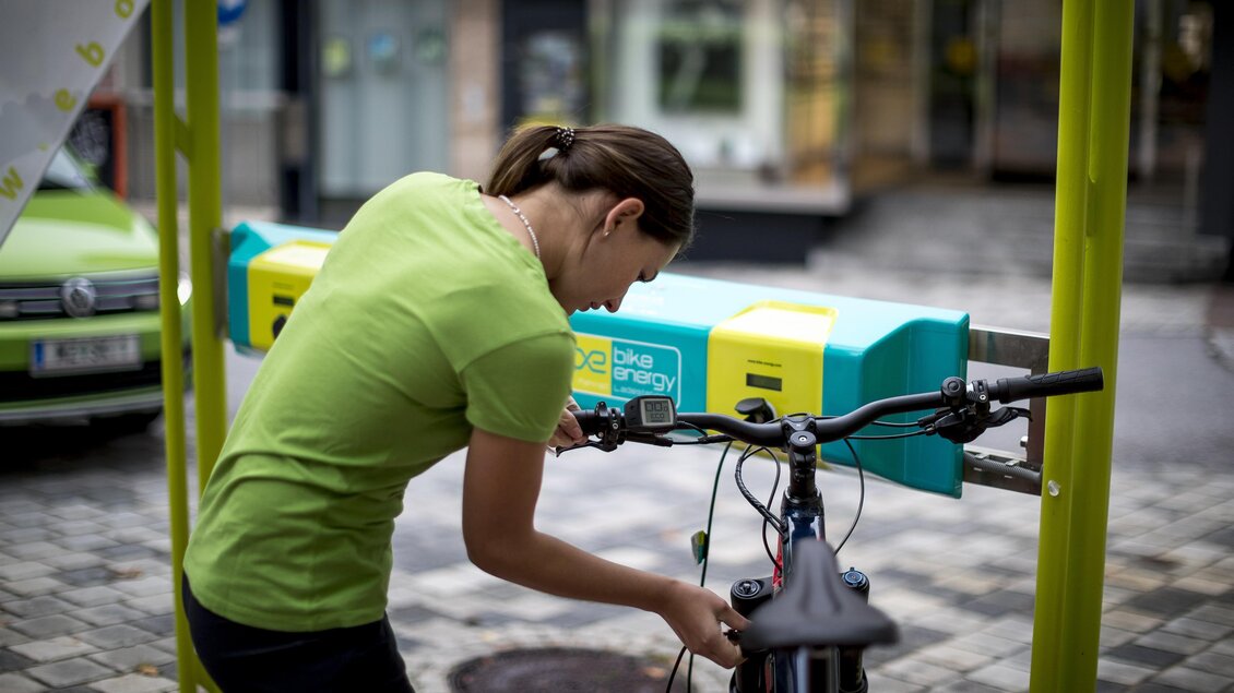 Eine Person in einem grünen T-Shirt repariert ein Fahrrad an einer Station. Im Hintergrund sind flache Gebäude und eine ruhige Straße zu sehen. | © TV Hochsteiermark