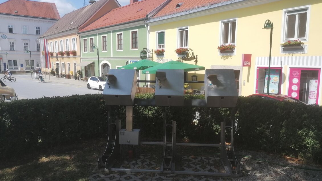 Drei moderne Fahrradrastplätze mit Verleihstation in einer hübschen Stadt. Bunte Häuser und ein klarer Himmel sind im Hintergrund sichtbar. | © Sabrina Prattes