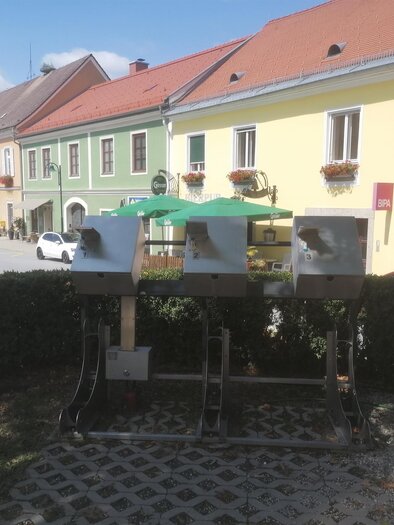 Three modern bike rest areas with rental stations in a nice town. Colorful houses and a clear sky are visible in the background. | © Sabrina Prattes