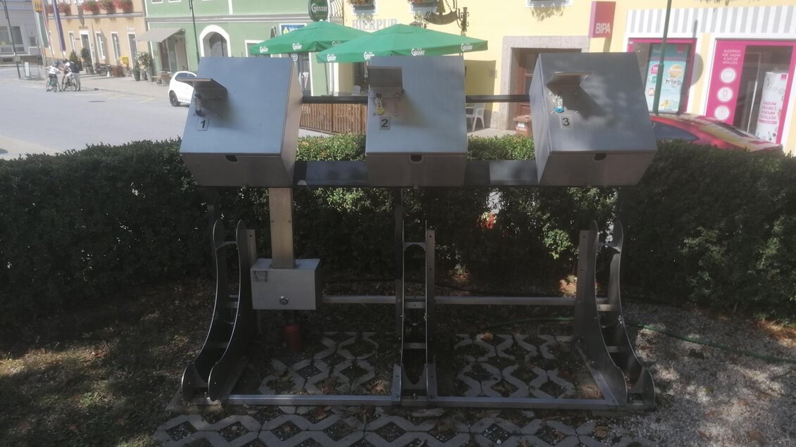 Drei silberne Abfallbehälter stehen auf einem Pflastersteinboden. Im Hintergrund sind bunte Gebäude und ein Sonnenschirm zu sehen. | © Sabrina Prattes