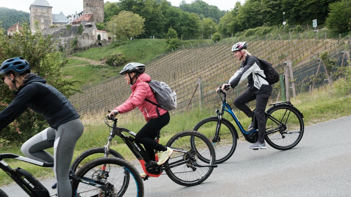 Eine Gruppe von Radfahrern fährt auf einem Weg entlang von Weinbergen. Im Hintergrund ist eine Burg und grüne Bäume zu sehen. | © Schilcherland Steiermark