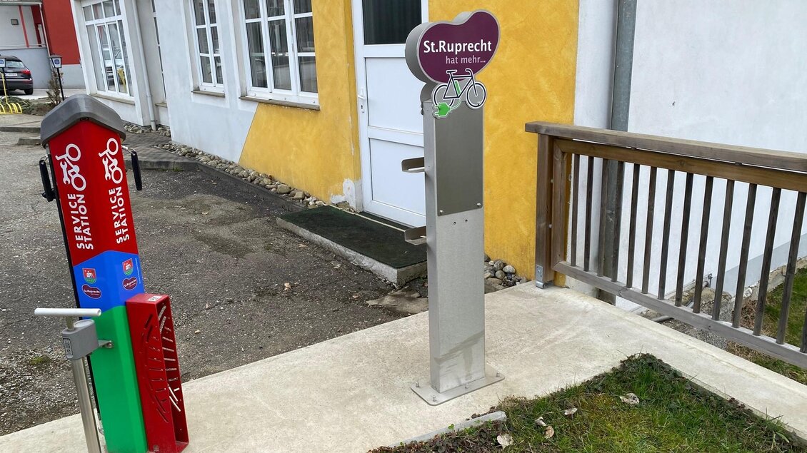 Eine Fahrradstation mit einem Reparaturset und einer Informationssäule in St. Ruprecht. Im Hintergrund sind bunte Gebäude und eine gemütliche Atmosphäre zu sehen. | © Oststeiermark Tourismus