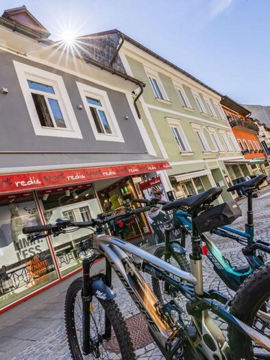 A lively street view with several bicycles parked. In the background, colorful buildings and a café can be seen. | © www.sportredia.at