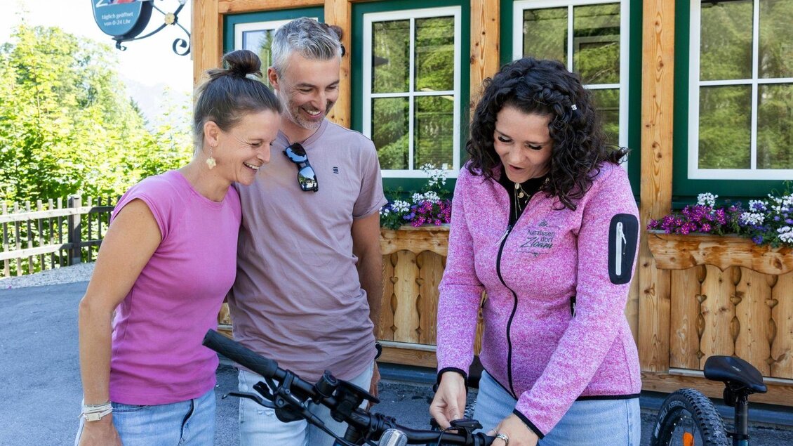Drei Personen stehen vor einem Holzhaus und schauen gemeinsam auf ein Fahrrad. Die Atmosphäre ist freundlich und entspannt. | © www.zloam.at