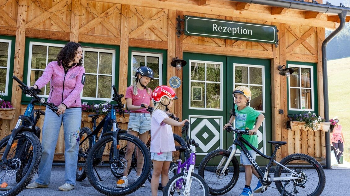 Eine Gruppe von Kindern und Erwachsenen steht vor einem Holzgebäude mit dem Schild "Rezeption". Alle tragen Helme und haben Fahrräder dabei. | © www.zloam.at