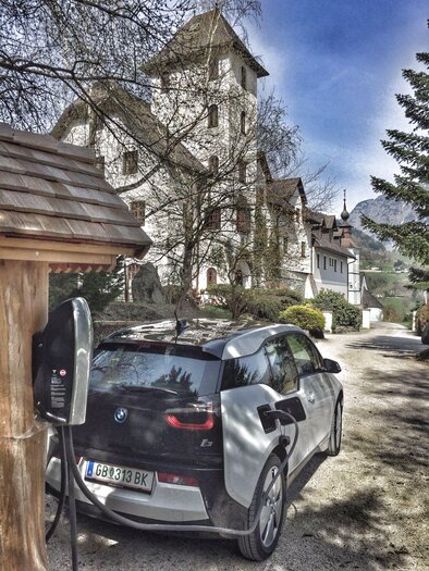 A modern electric car is parked at a charging station in front of a historic building. The surroundings are green and calm, with trees and a clear sky.
