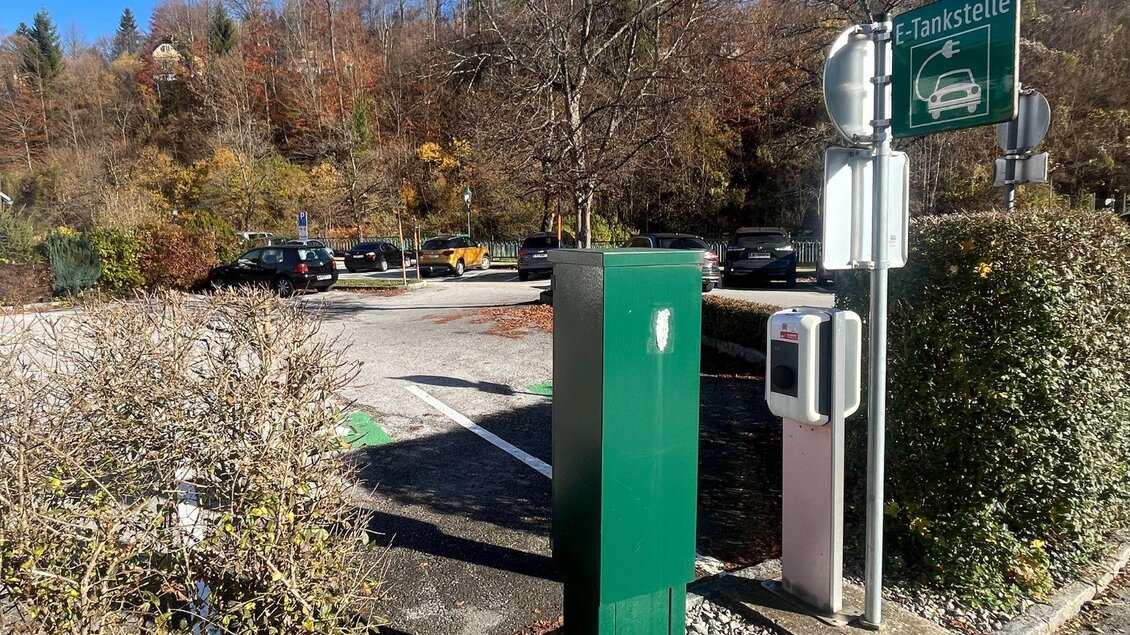 Eine Elektro-Tankstelle mit einer grünen Ladestation in einem Parkplatz. Im Hintergrund sind parkende Autos und Herbstbäume zu sehen. | © TVB Ausseerland- Salzkammergut_Nathalie Schröttenhamer