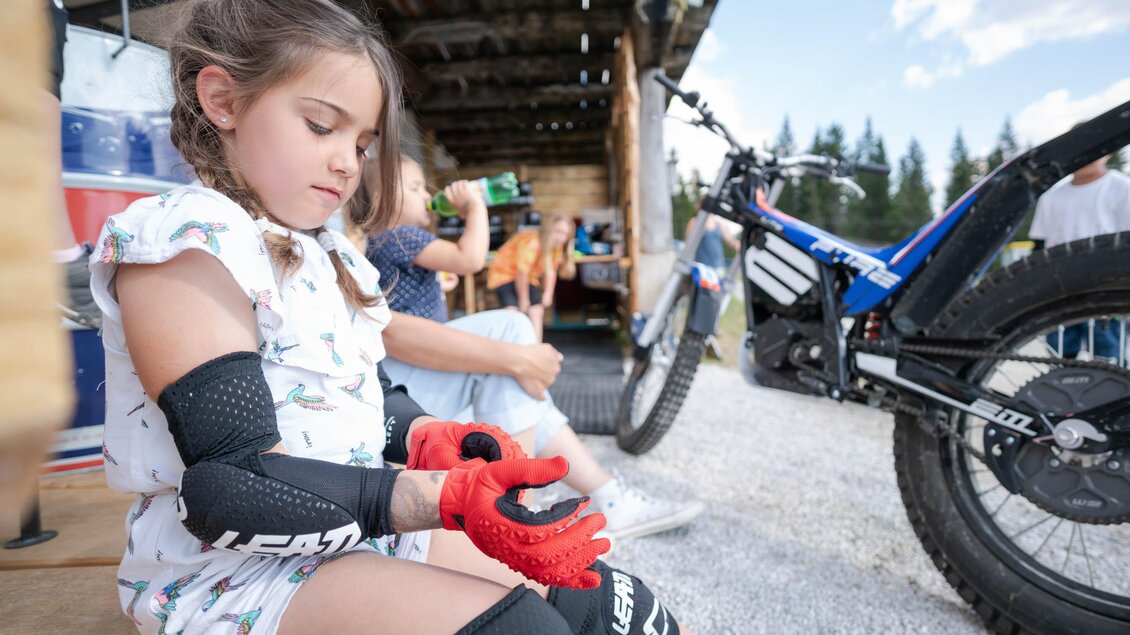 Ein Mädchen zieht Schutzkleidung an, während sie sich auf eine Fahrt mit ihrem E-Bike vorbereitet. Im Hintergrund sind andere Menschen und eine hölzerne Struktur zu sehen. | © Trialstars - Andreas Pilz