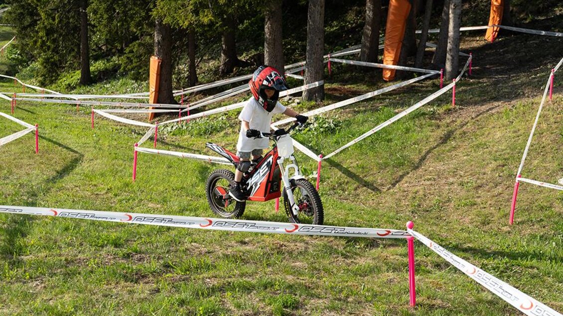 Ein Kind fährt auf einem kleinen Motorrad auf einem Parcours im Freien. Umgeben von Bäumen und Markierungsbändern zeigt die Szene einen spannenden Motocross-Aktivität. | © Trialstars - Josh Absenger