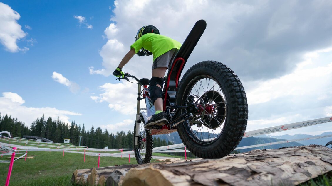 Ein Junge fährt mit einem Fahrrad über einen Baumstamm. Der Himmel ist blau mit einigen Wolken und im Hintergrund sind Bäume sichtbar. | © Trialstars - Andreas Pilz