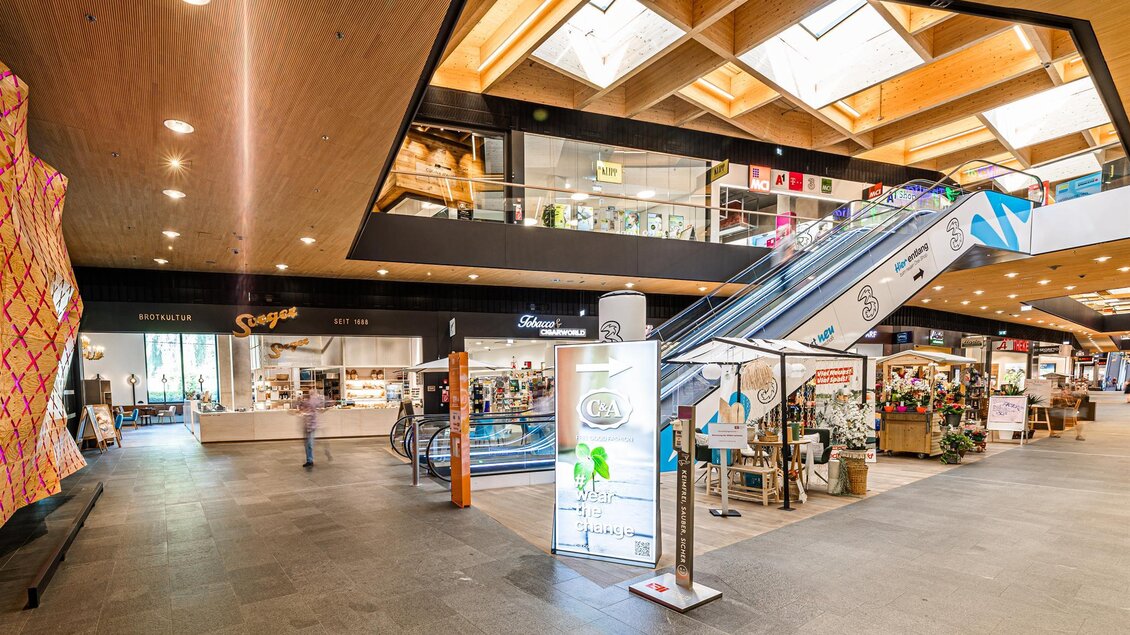 Ein modernes Einkaufszentrum mit einem offenen Raum und einer eleganten Holzdecke. Im Vordergrund sieht man eine Rolltreppe und verschiedene Geschäfte. | © Thomas Sattler