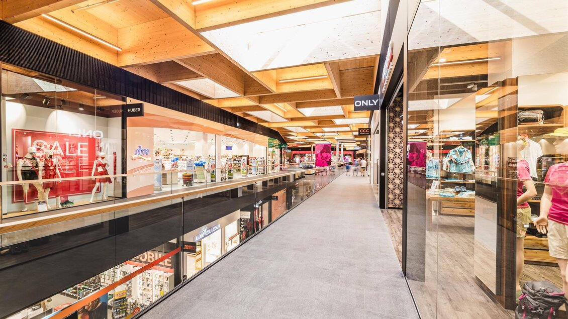 Ein moderner Einkaufsbereich mit hellen Decken und Glasgeländern. Verschiedene Geschäfte sind links und rechts zu sehen, die eine Vielzahl von Produkten anbieten. | © Thomas Sattler