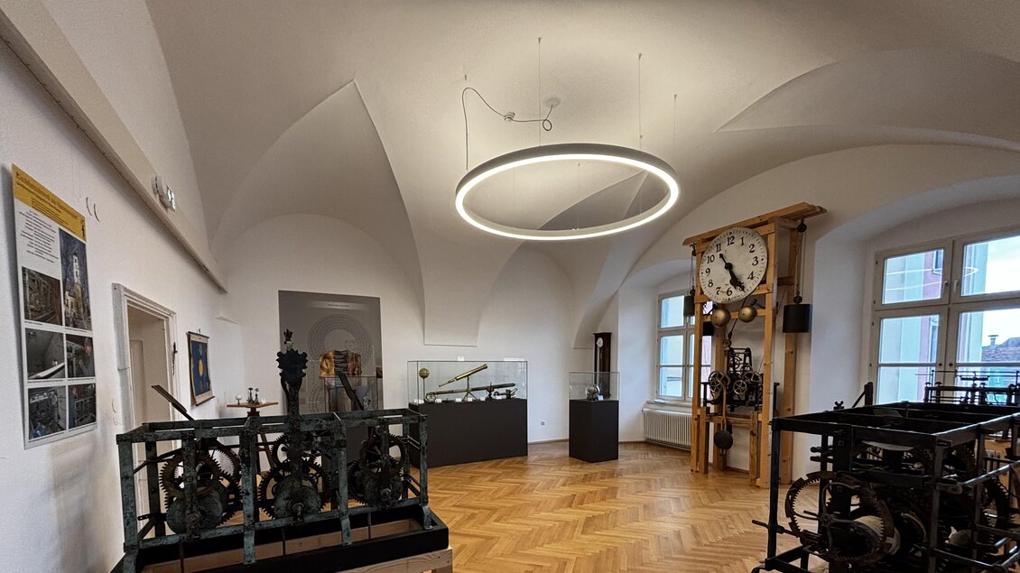 Ein moderner Ausstellungsraum mit einer runden Deckenlampe. Im Raum befinden sich historische Geräte und eine große Uhr. | © Echophysics