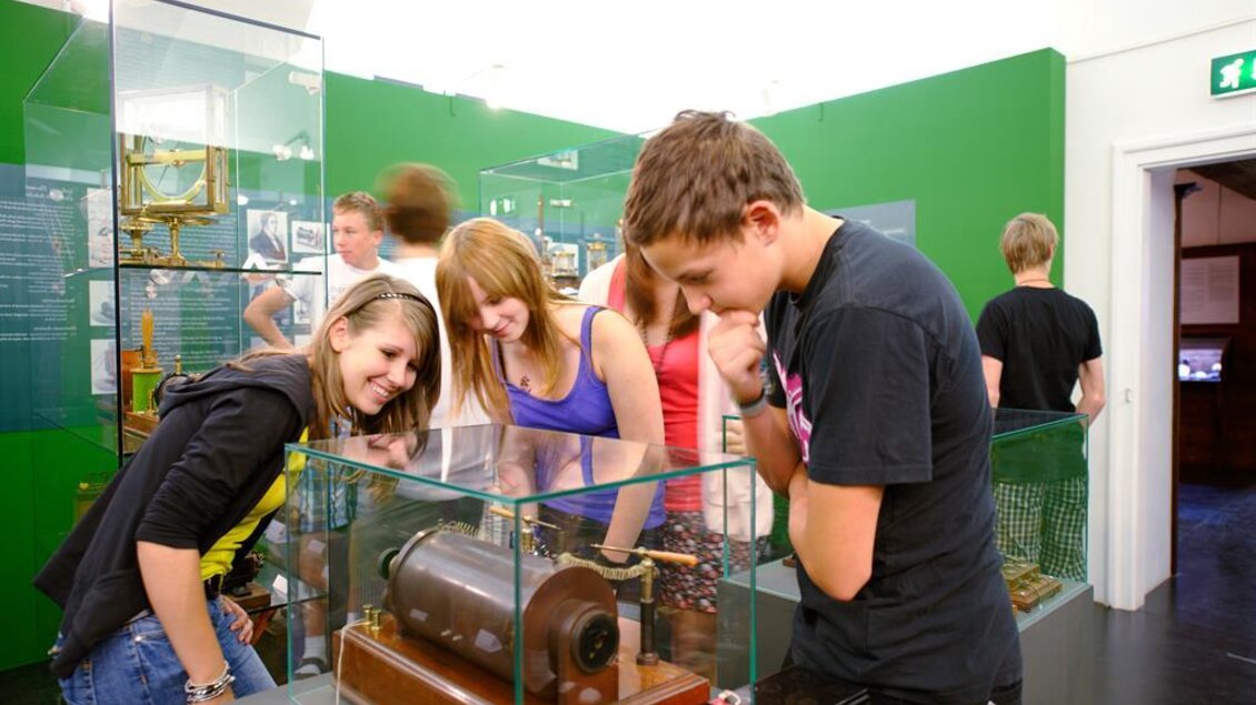 Eine Gruppe von Jugendlichen betrachtet neugierig ein Ausstellungsstück in einem Museum. Im Hintergrund sind weitere Besucher und Vitrinen mit Exponaten zu sehen. | © Peter Schuster