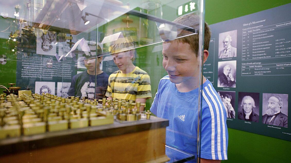 Zwei Jungen stehen vor einem Vitrinenausstellung, die historische Objekte zeigt. Im Hintergrund sind informierende Tafeln und Porträts von bekannten Persönlichkeiten zu sehen. | © Peter Schuster