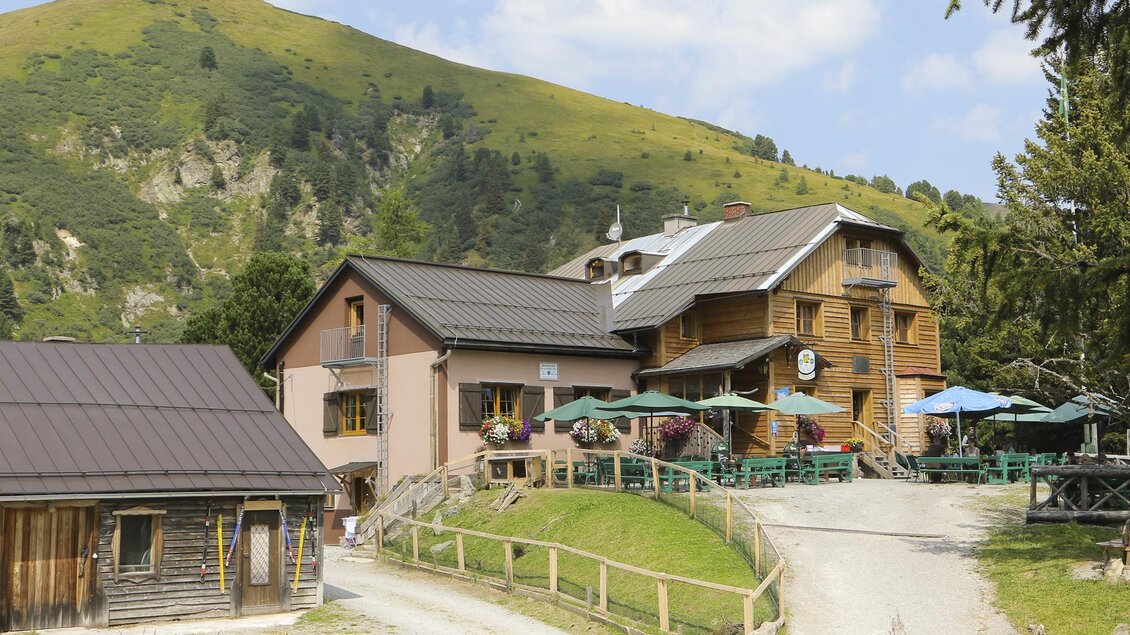 Ein charmantes Berghaus mit einer Holzfassade und Terrassen. Im Hintergrund sieht man eine grüne Hügellandschaft unter einem blauen Himmel. | © Erlebnisregion Murtal