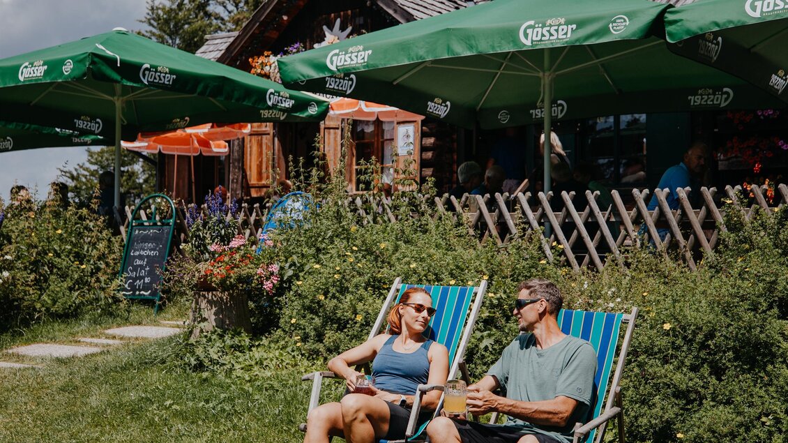 Ein Paar sitzt entspannt in Liegestühlen vor einem rustikalen Gebäude. Im Hintergrund sind grüne Sonnenschirme zu sehen. | © nicoleseiser.at