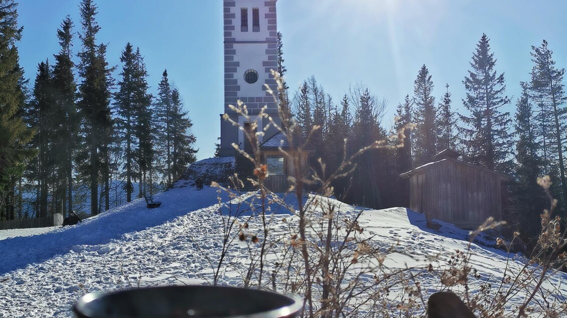 Eine schneebedeckte Landschaft mit einem hohen, weißen Turm im Hintergrund. Im Vordergrund steht eine Tasse. | © TV Hochsteiermark /  Brigitte Digruber