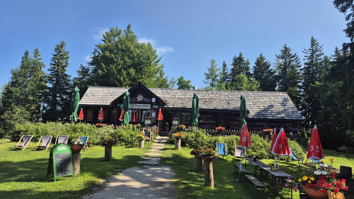 Ein gemütliches Restaurant in der Natur mit Gartenmöbeln und Sonnenschirmen. Im Hintergrund stehen hohe Bäume und ein klarer, blauer Himmel.