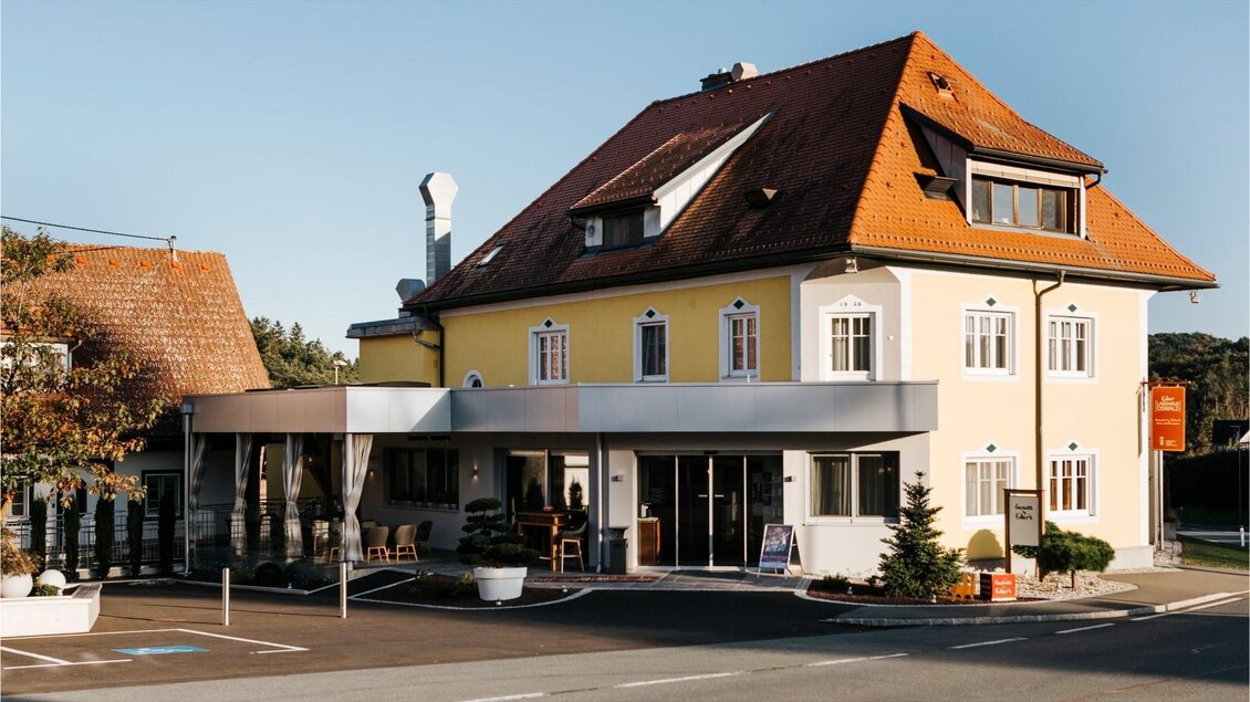 Ein charmantes Gebäude mit hellgelber Fassade und roten Ziegeldächern. Im Vordergrund ist ein moderner Eingangsbereich mit Sitzgelegenheiten. | © Edler Landhaus Oswald